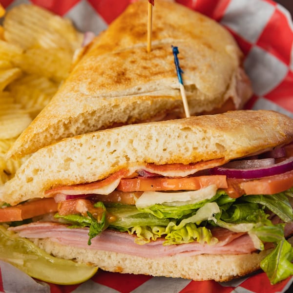 A Grace's Place Pizzeria Italian sub sandwich with chips and a pickle spear in a red checkered basket.